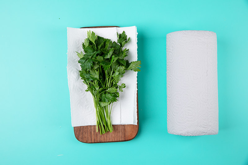 A bunch of herbs laying on a paper towel on a cutting board. A roll of paper towels lays to the right of it.