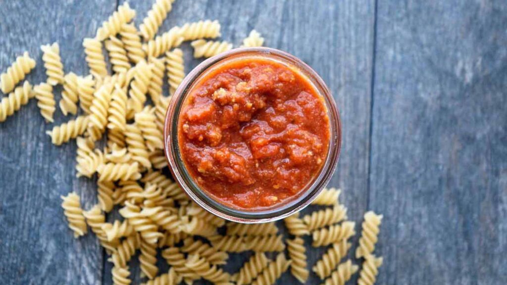 And overhead view of this homemade spaghetti sauce looking down into the open jar of sauce. Dry spiral pasta lays on the table at the base of the jar.