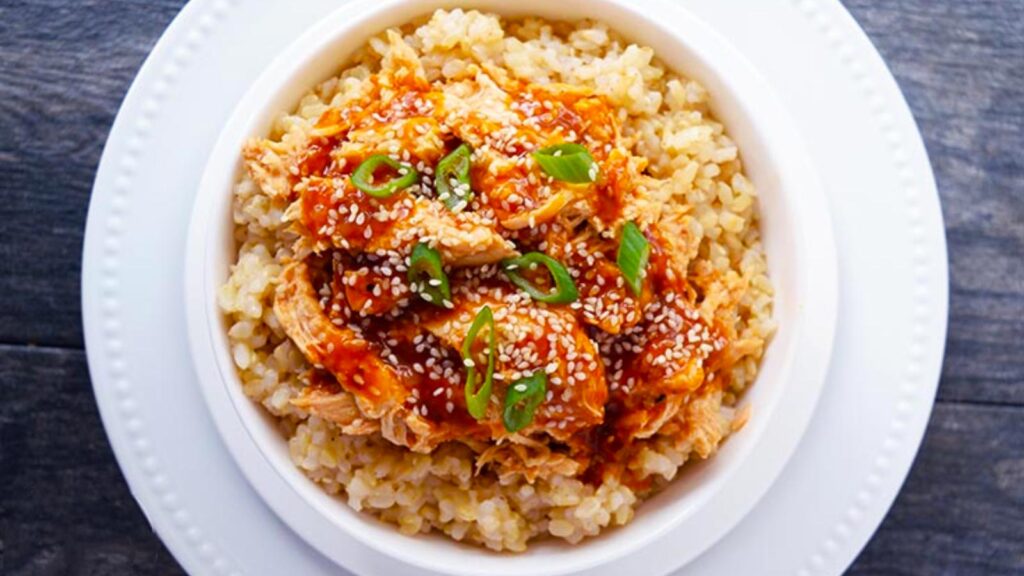 An overhead view of a white bowl filled with rice and topped with honey sesame chicken.