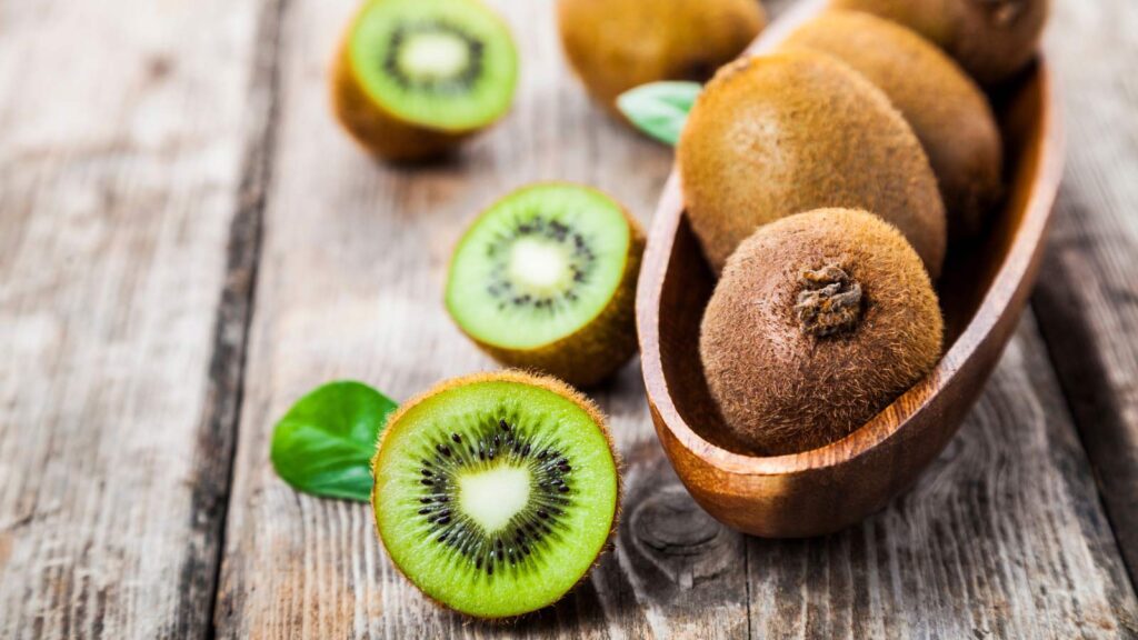 Kiwi fruit in a bowl with a few cut kiwi halves laying on a wood table next to the bowl.