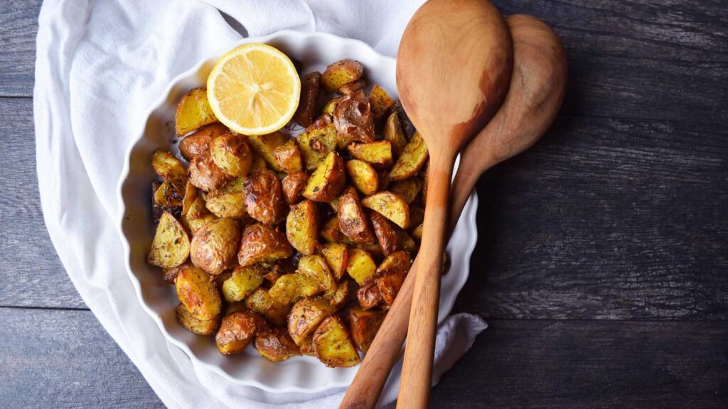 An overhead view of a white, fluted dish filled with lemon garlic potatoes. A wedge sits in the side of the dish. Wood spoons rest over the other side of the dish.