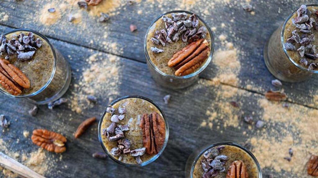Individual cups of Maca Chia Seed Pudding on a table with maca powder and pecans sprinkled around them.