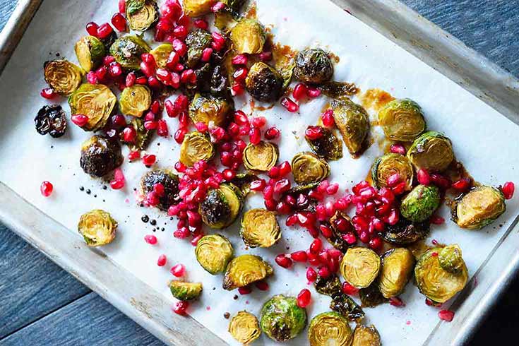 Maple Roasted Brussels Sprouts laying on a parchment-lined baking sheet with pomegranate arils sprinkled over them.
