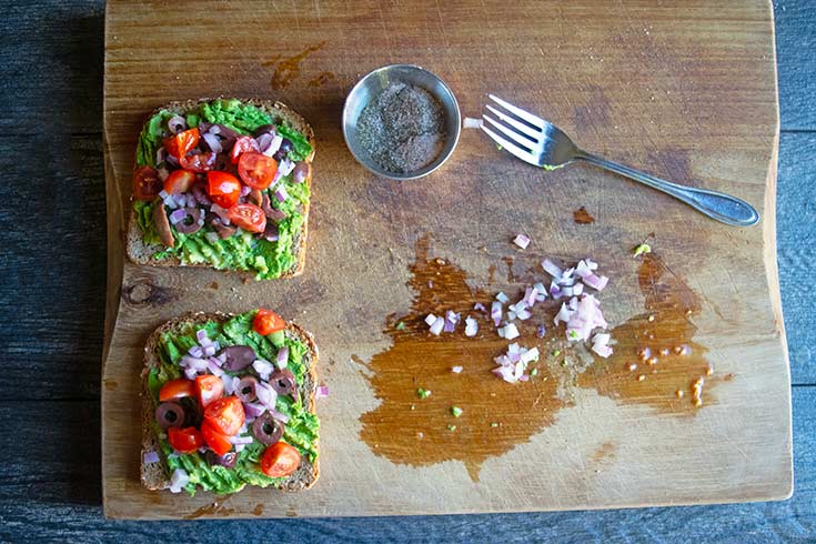 Mediterranean toppings added to avocado toast on a cutting board.
