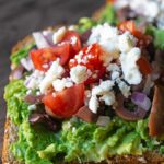 A side view of two slices of Mediterranean Avocado Toast on a cutting board.