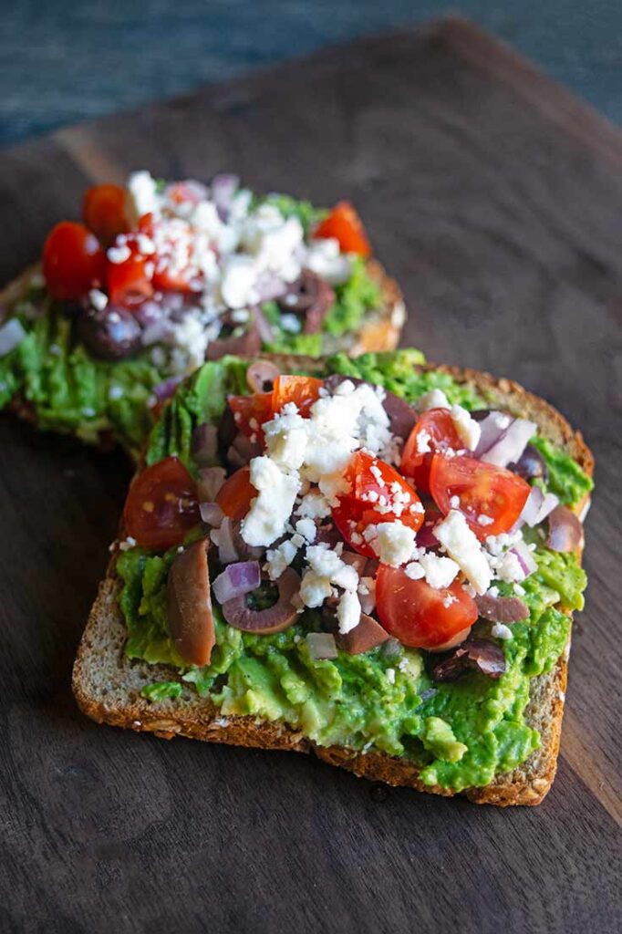 A front view of two slices of Mediterranean Avocado Toast on a cutting board.