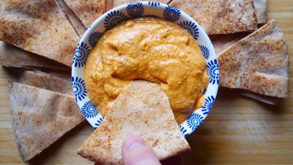 A hand dips a pita triangle into a bowl of dip.