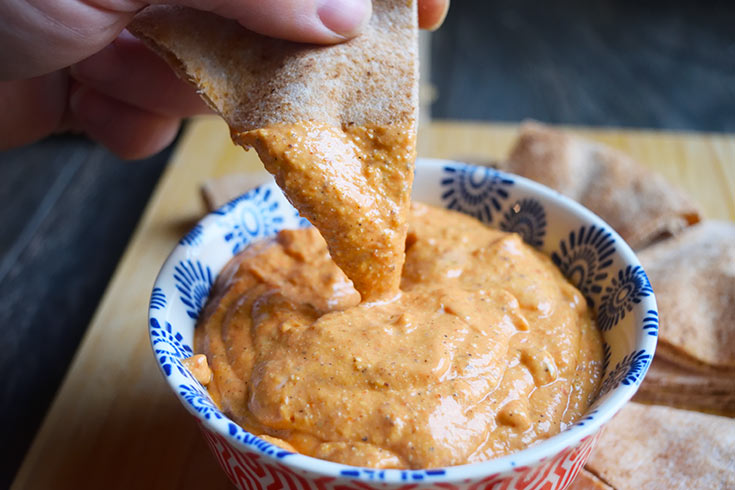 A closeup of a hand dipping a pita bread triangle into a bowl of Mediterranean Roasted Red Pepper Dip.
