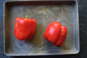 Two fresh, raw, red bell peppers laying on a sheet pan.