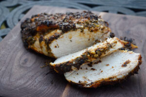 A finished Herb-Infused Mediterranean Turkey Breast laying on a cutting board with two slices laying at the front of the breast.