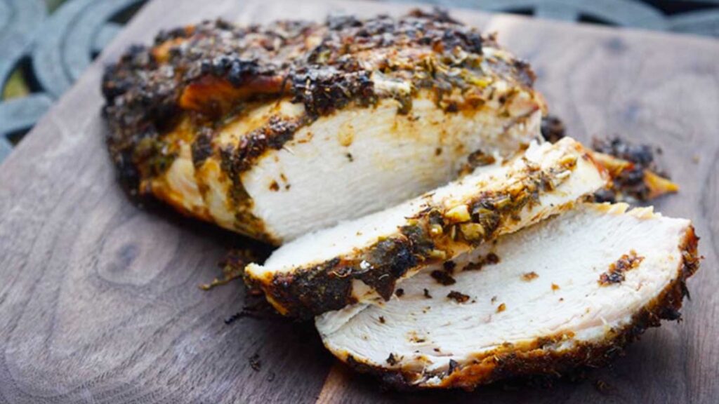 A finished Herb-Infused Mediterranean Turkey Breast laying on a cutting board with two slices laying at the front of the breast.