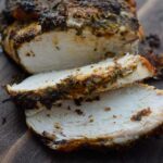 A front view of a Herb-Infused Mediterranean Turkey Breast laying on a cutting board with two slices carved.
