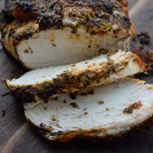 A front view of a Herb-Infused Mediterranean Turkey Breast laying on a cutting board with two slices carved.