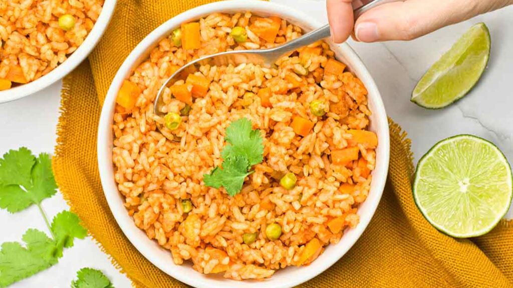 A hand spoons some Mexican rice out of a white bowl full of it.