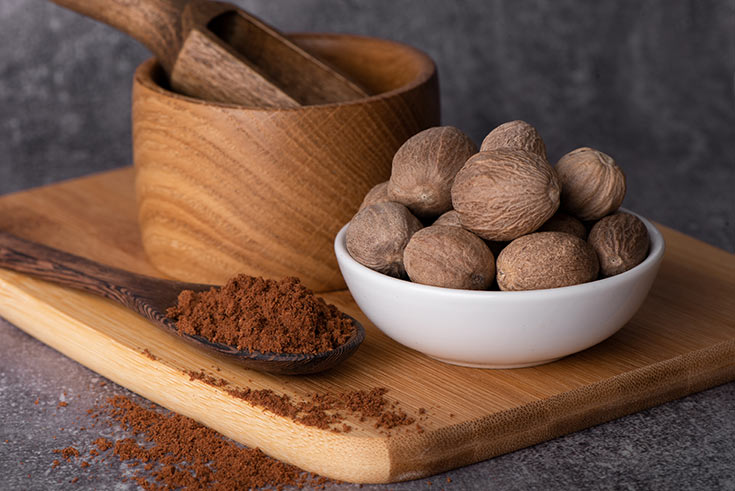 A bowl of nutmegs sits next to a spoon full of ground nutmeg.