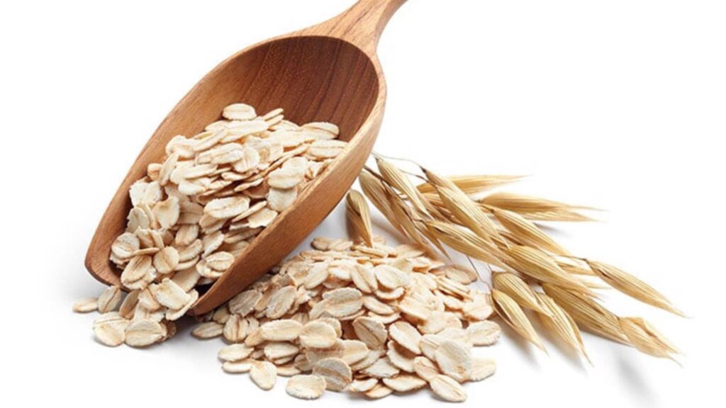 A wooden scoop sits filled with oats next to a pile of oats on a white background.