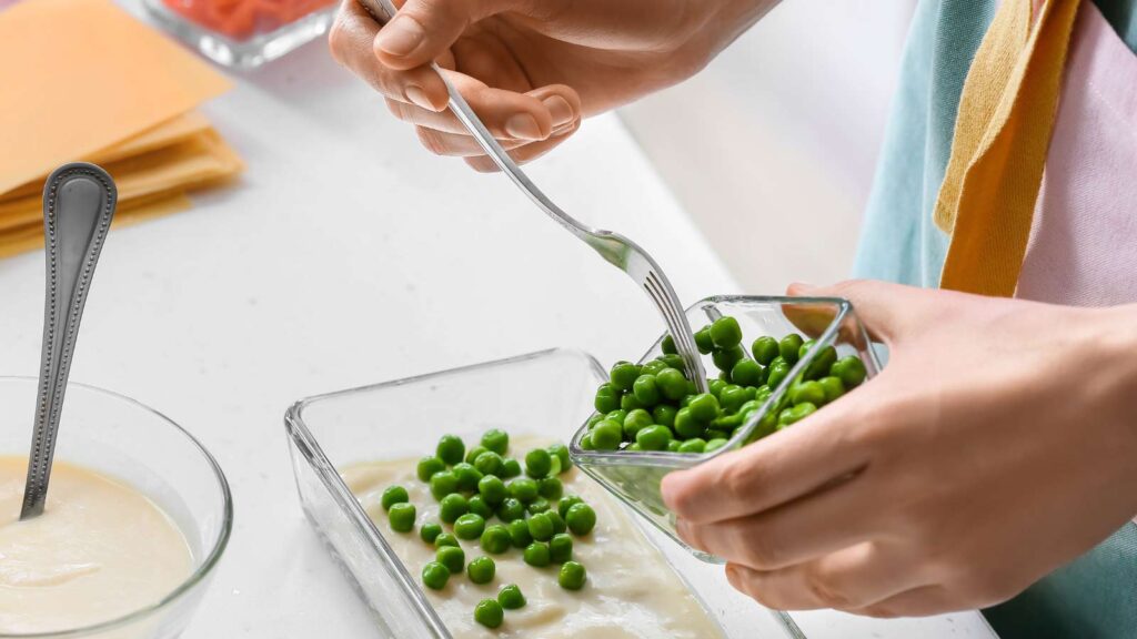 A woman adding peas to a layer of lasagna.
