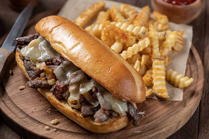 A Philly cheesesteak on a wood cutting board with a pile of french fries next to it.
