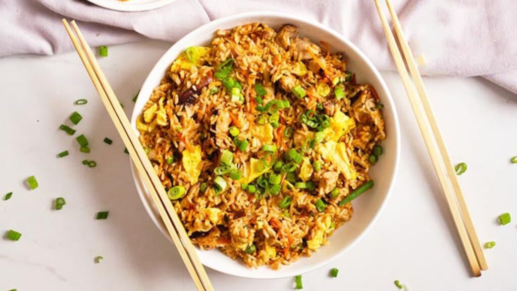 A white bowl filled with pork fried rice. Chopstick lay on the bowl and to the right of it.