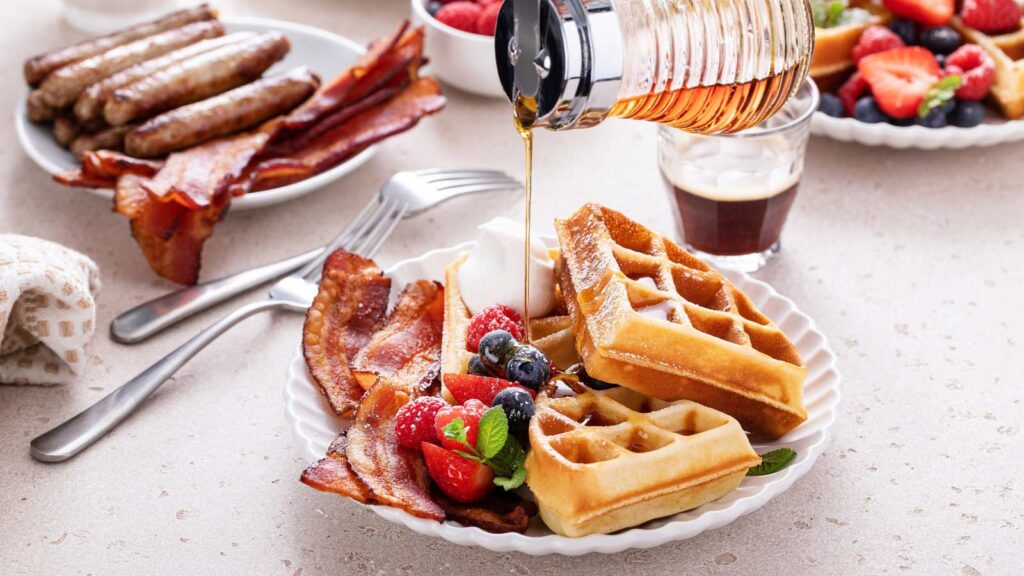 Maple syrup being poured over a plate with waffles, berries and bacon.