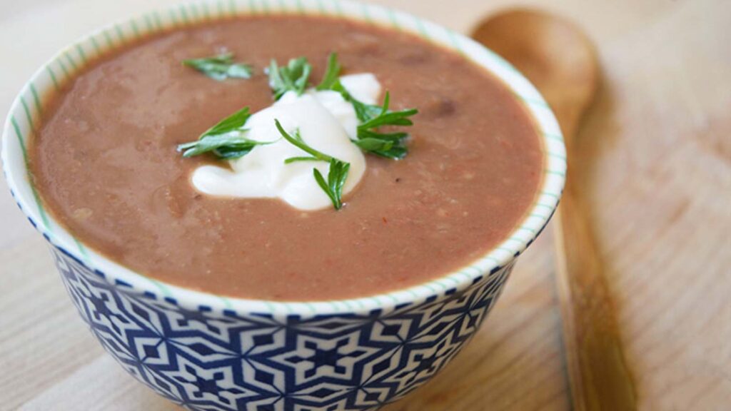 A small bowl of Refried Bean Soup on a wood table. A wood spoon lays next to the bowl.