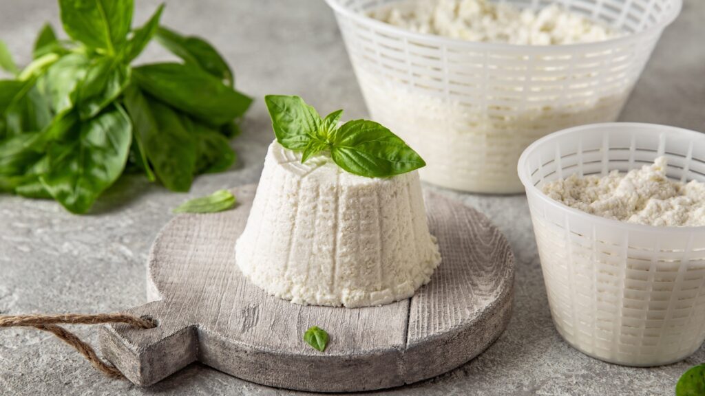 A formed mound of ricotta cheese on a cutting board next to two baskets of more ricotta cheese.