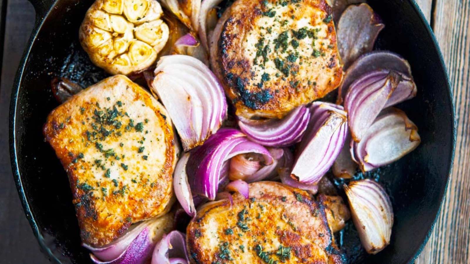 An overhead view of a cast iron skillet filled with three browned pork chops, chunks of red onion and a whole head of garlic.