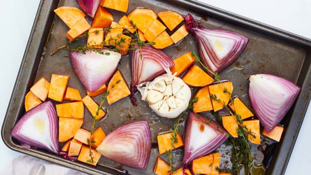 The onions, garlic and sweet potatoes with herbs and oil, sitting on a baking sheet.
