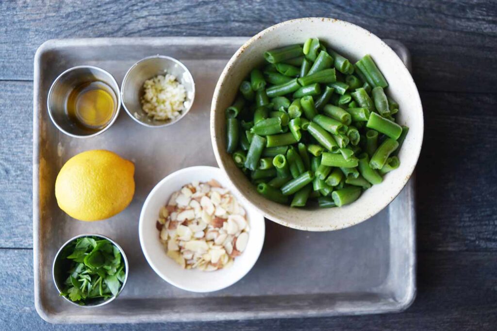 Sautéed Green Beans With Almonds Recipe ingredients in individual bowls sitting on a sheet pan.
