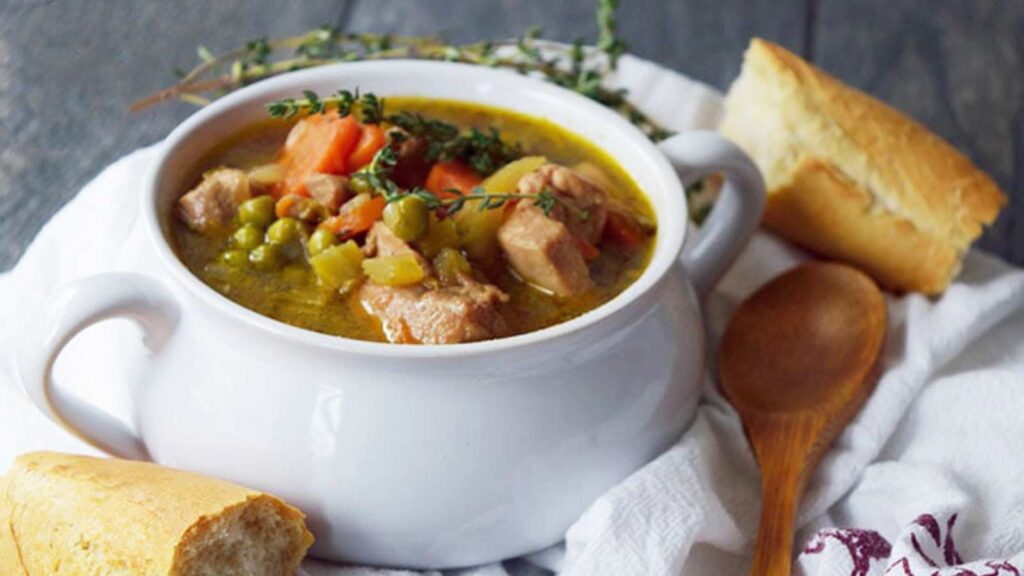 A white bowl filled with Slow Cooker Chicken Stew, surrounded by pieces of baguette and a wooden spoon.