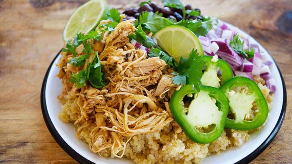 A plate holds some quinoa piled high with shredded cilantro lime chicken with a side of black beans, chopped red onions, sliced jalapeno peppers, and fresh lime wedges.