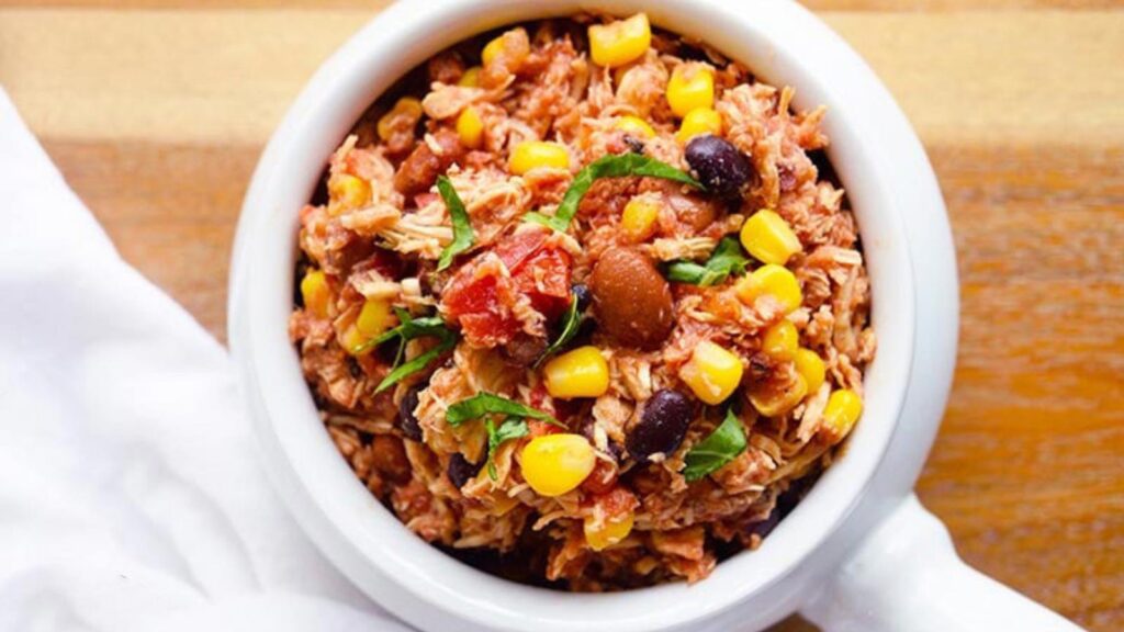 A white crock filled with Slow Cooker Southwestern Chicken, sits on a wood surface next to a white towel.