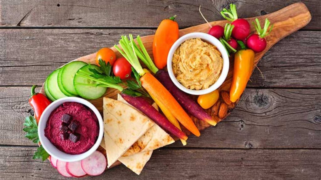 An overhead view of a charcuterie board with veggies, pita chips, and two bowls of dip.