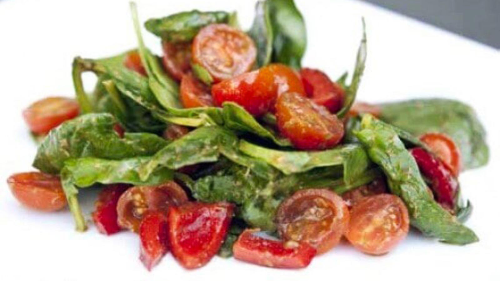 Spinach salad with pesto dressing and halved cherry tomatoes on a white plate.