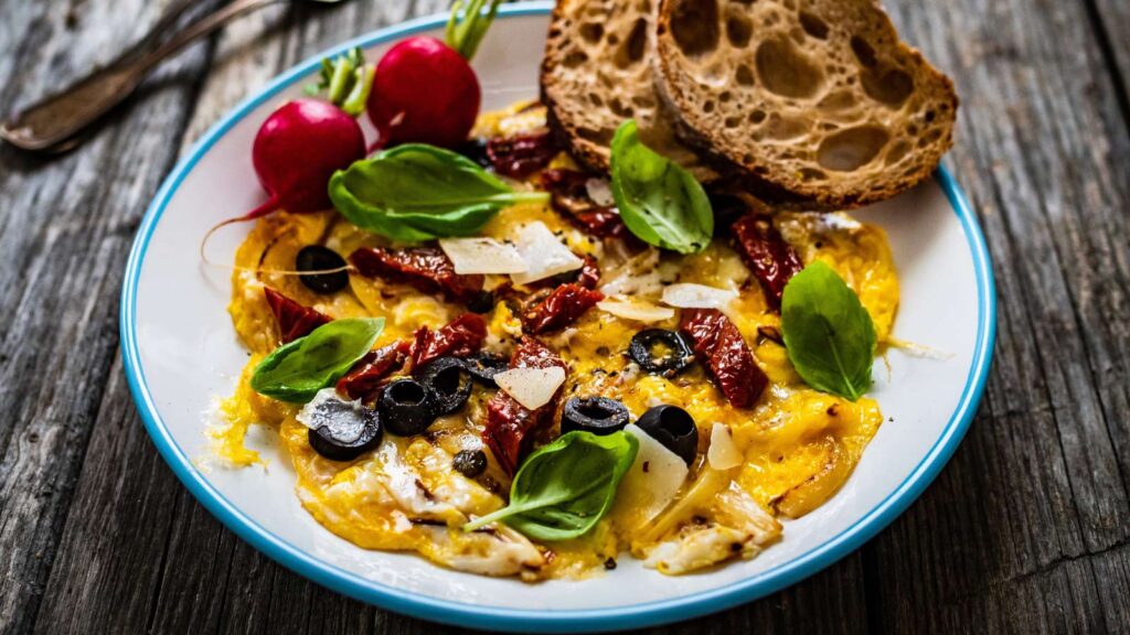 Delicious breakfast - scrambled eggs with onion, sun dried tomato, basil, parmesan and black olives on wooden table.