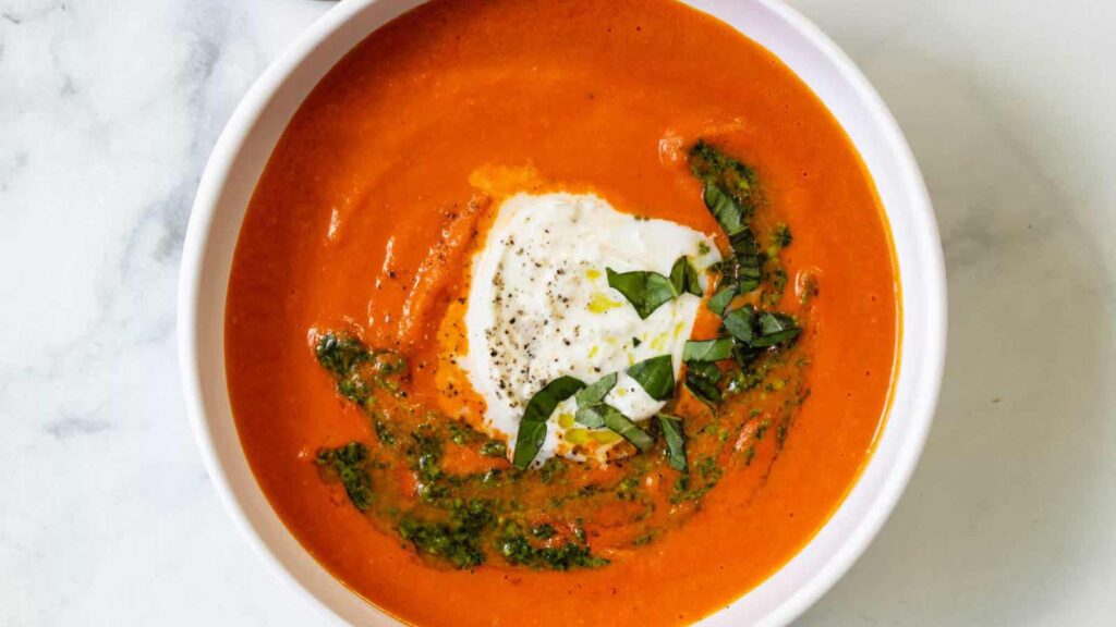A white bowl filled with sweet potato red pepper soup and garnished with cream and fresh basil.