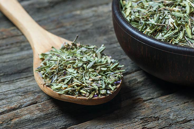 A bowl and wooden spoon sit on a wooden table and are filled with dried thyme leaves.