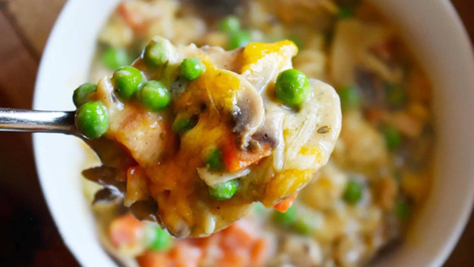 A bowl filled with tuna noodle casserole sits on a table as a spoon lifts a spoonful of it up and out of the bowl.