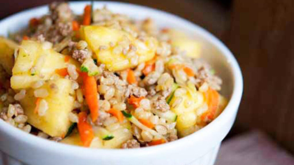 A white bowl sits on a wood surface filled with curry pineapple turkey rice bowl.