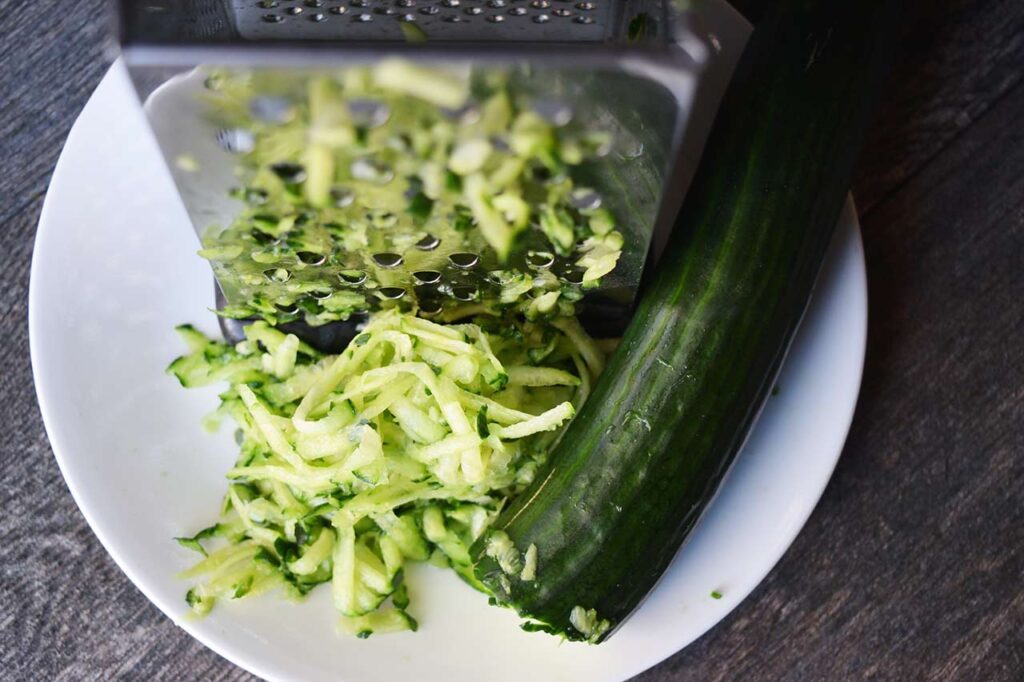 A partially grated English cucumber lays on a plate next to a grater and a partially whole cucumber.