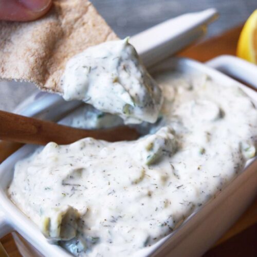 A pita chip with Tzatziki sauce on it over a dish filled with the dip.