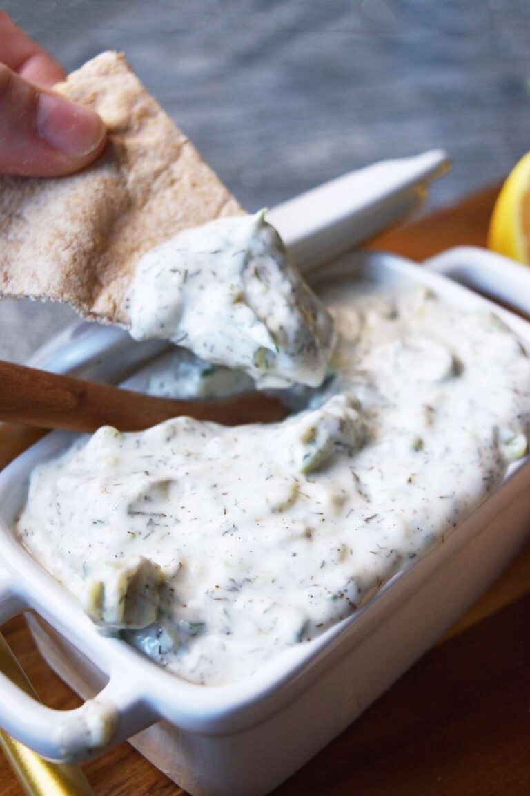 A pita chip with Tzatziki sauce on it over a dish filled with the dip.