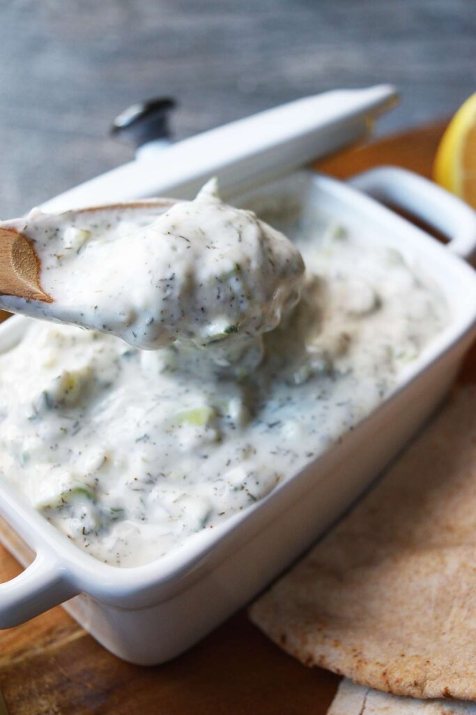A wood spoon lifts a spoonful of Tzatziki out of a white dish that is filled with it.