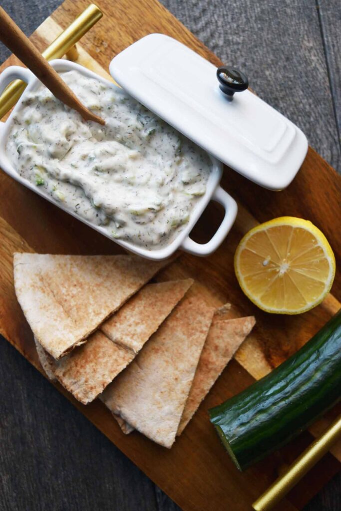 An overhead view of a cutting board holding a white dish filled with Tzatziki, pita chips, half a lemon and a cucumber.