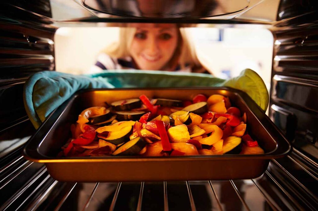 Woman Putting Dish Of Vegetables Into Oven To Roast.