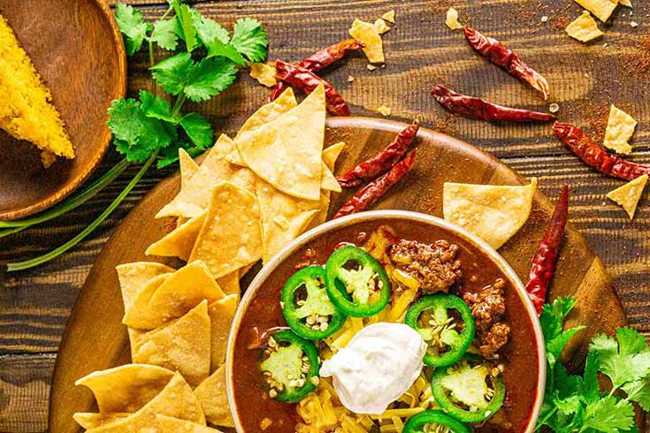 An overhead view of a bowl of venison chili surrounded by corn chips and chili peppers.