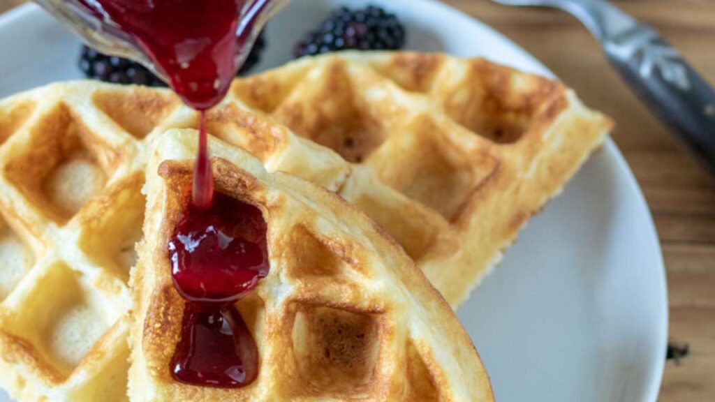 Berry syrup being poured over waffles on a plate.