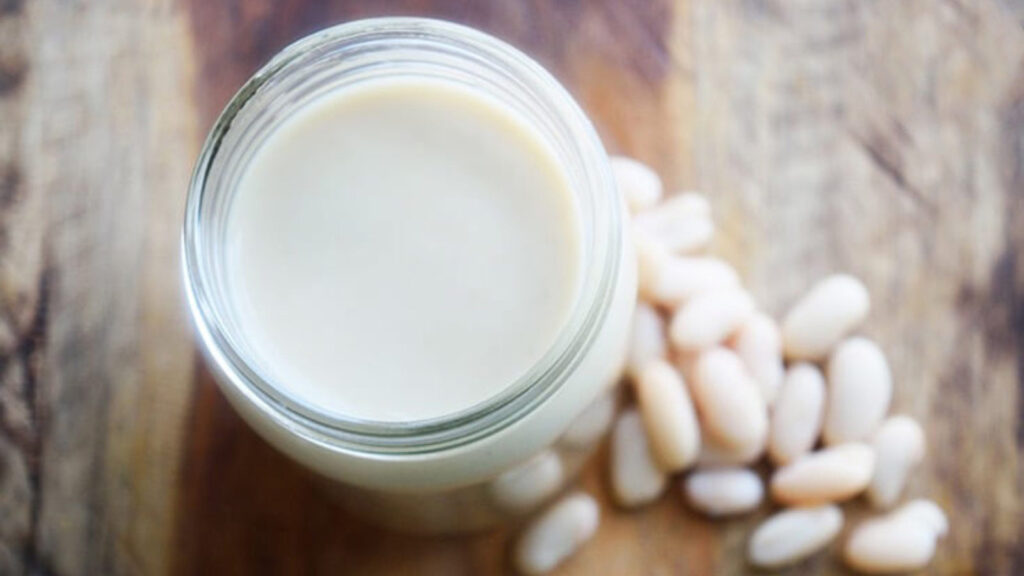 A jar filled with White Bean Alfredo Sauce with a few white beans laying in front of the jar on a wooden surface.