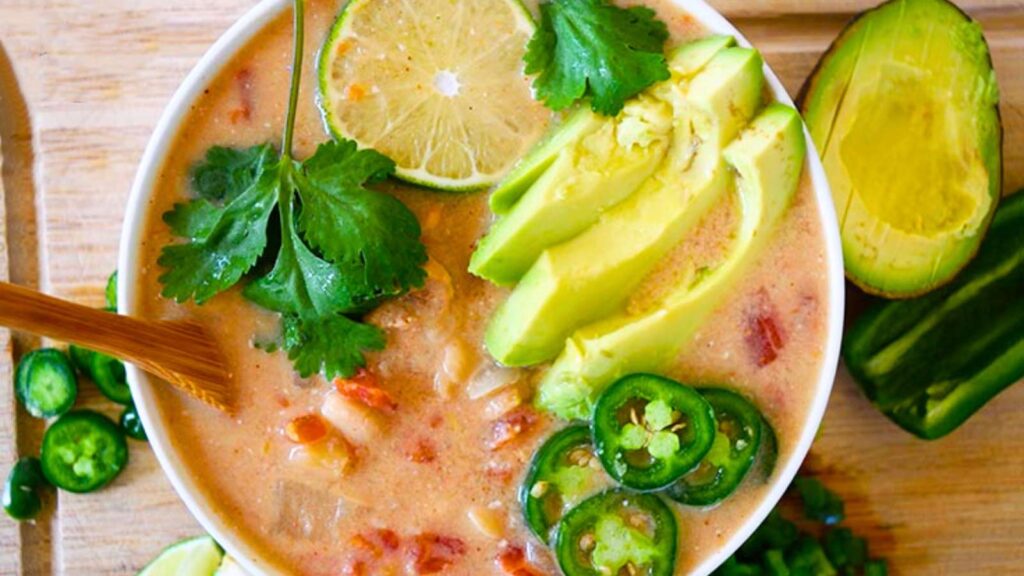 An overhead view of a bowl of white chicken chili that is garnished with avocado slices, a lime slice, jalapeno slices and fresh cilantro.