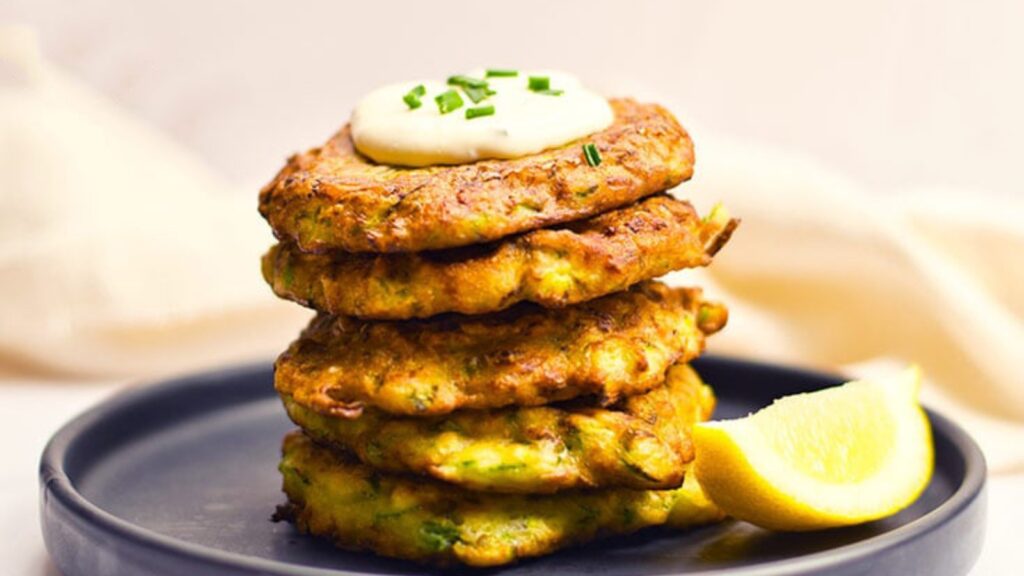 A stack of zucchini fritters on a plate, topped with a dollop of hummus on top.
